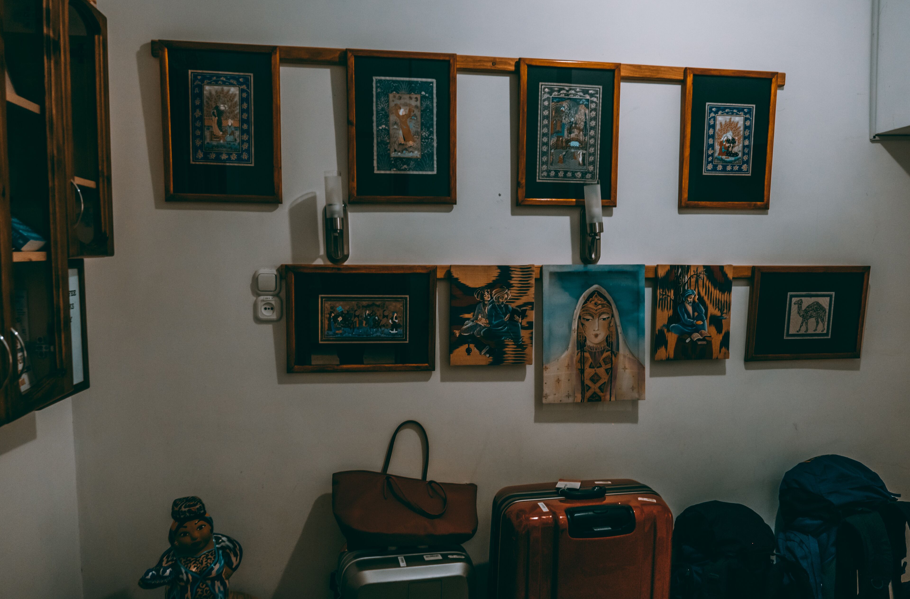 A room with a wall decorated with framed artwork, a wooden shelf, and a statue. There are suitcases and a backpack on the floor.