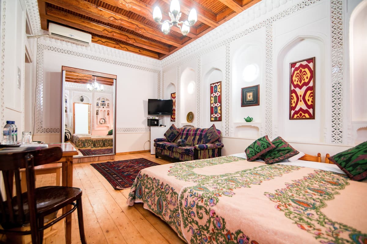 Ein traditionell eingerichteter Raum mit Holzbalkendecke, einem Bett mit blumigem Überwurf, einer Couch mit bunten Kissen, einem Fernseher und einem dekorativen Spiegel.