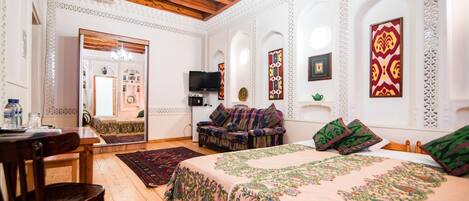Une chambre à l’ancienne, avec un plafond en bois, un lit recouvert d’une courtepointe à motifs floraux, un canapé agrémenté de coussins colorés, une télévision et un miroir décoratif.