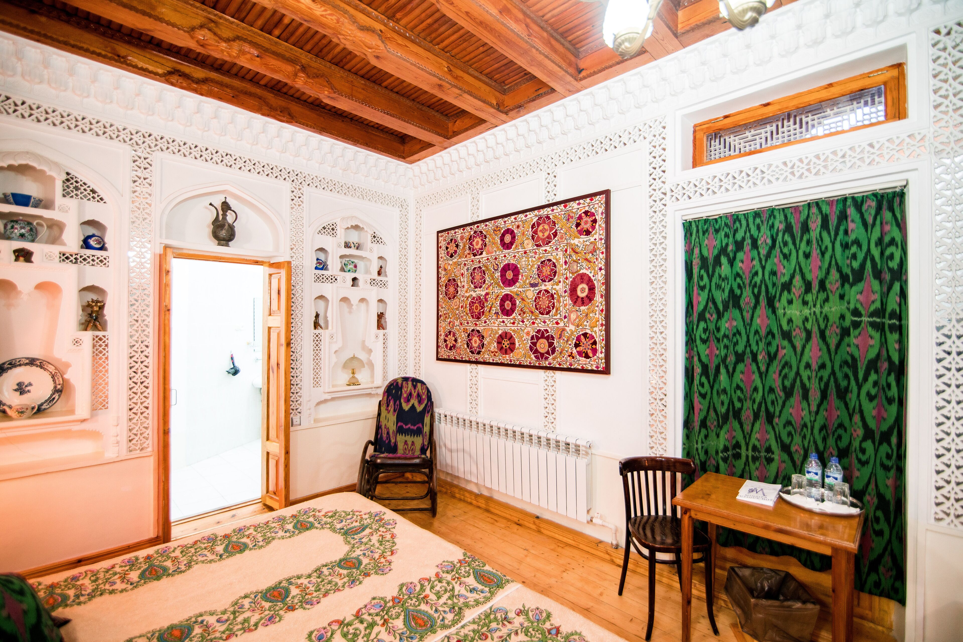 Una habitación con techo de madera, paredes blancas con diseños intrincados, una alfombra estampada, una mesa de madera con una bandeja y una silla.