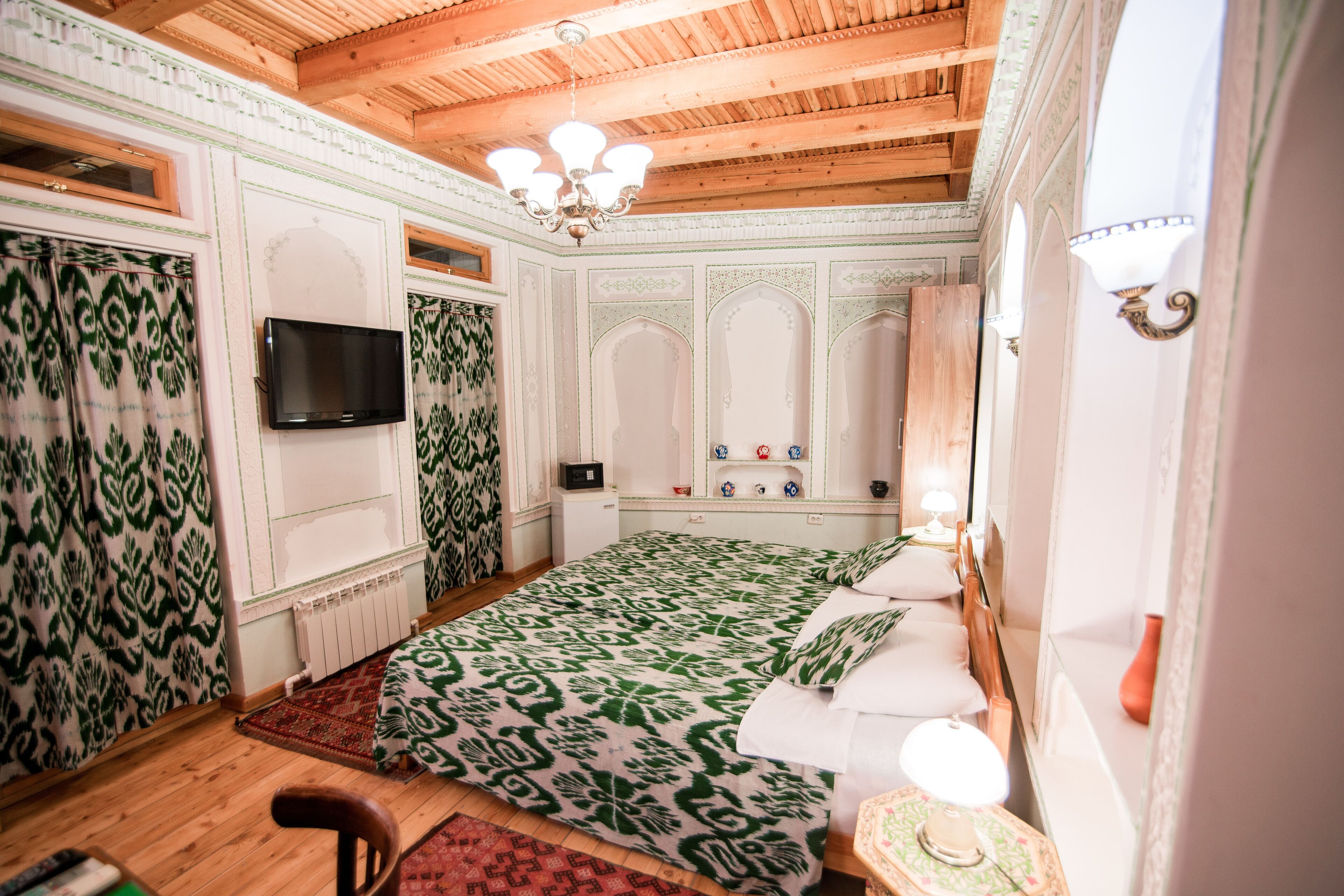 A room with a wooden ceiling, a bed with patterned bedding, a television, and a small table with a lamp.