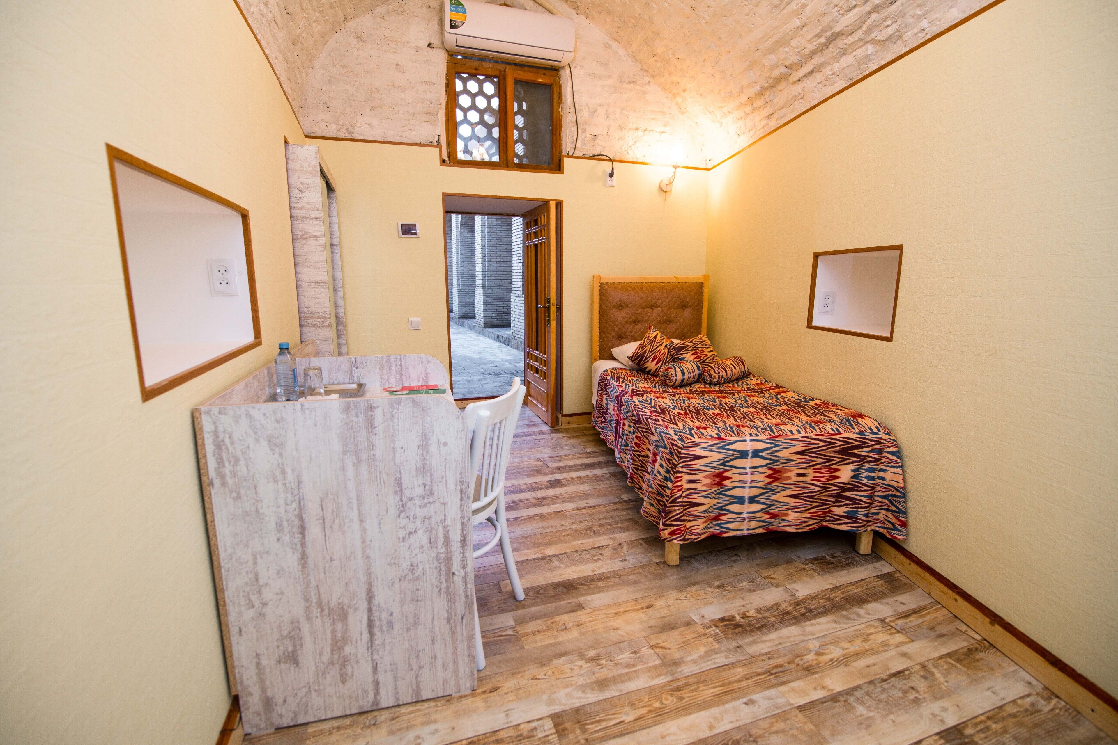 A small, cozy room with a bed, a desk, and a chair. There is a window with a view of the outside, and a door leading to another room.