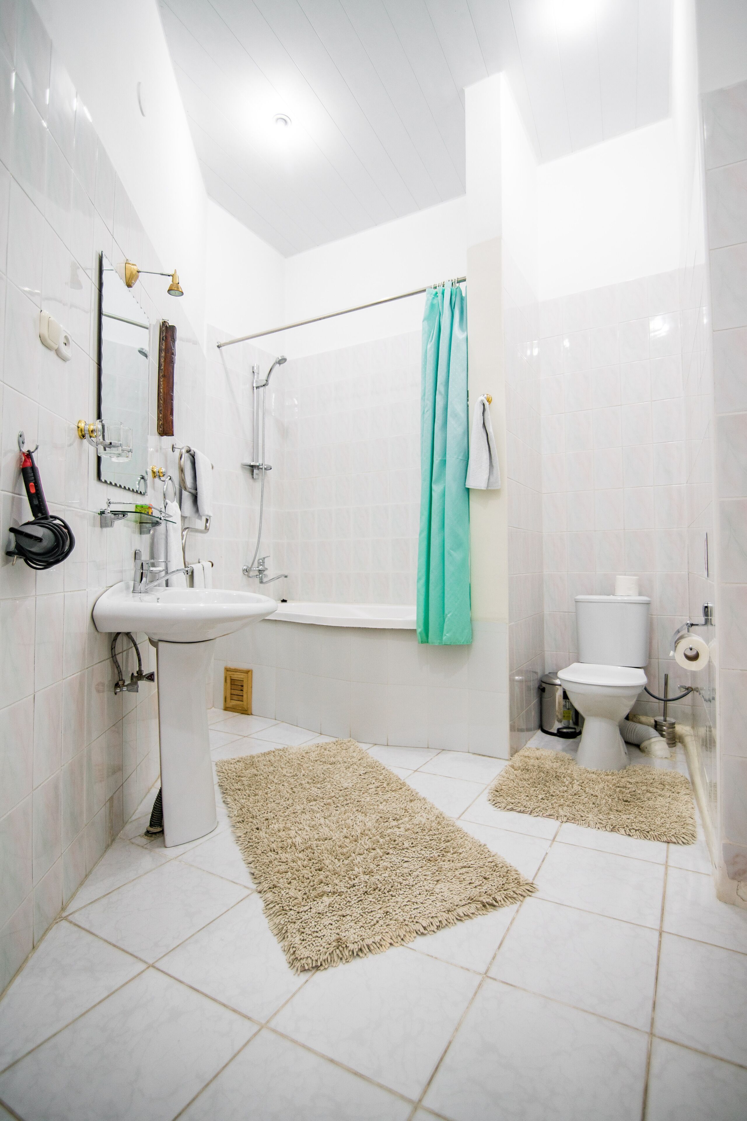 A modern bathroom with a white sink, bathtub, and toilet. There are two beige rugs on the floor, and a teal shower curtain.