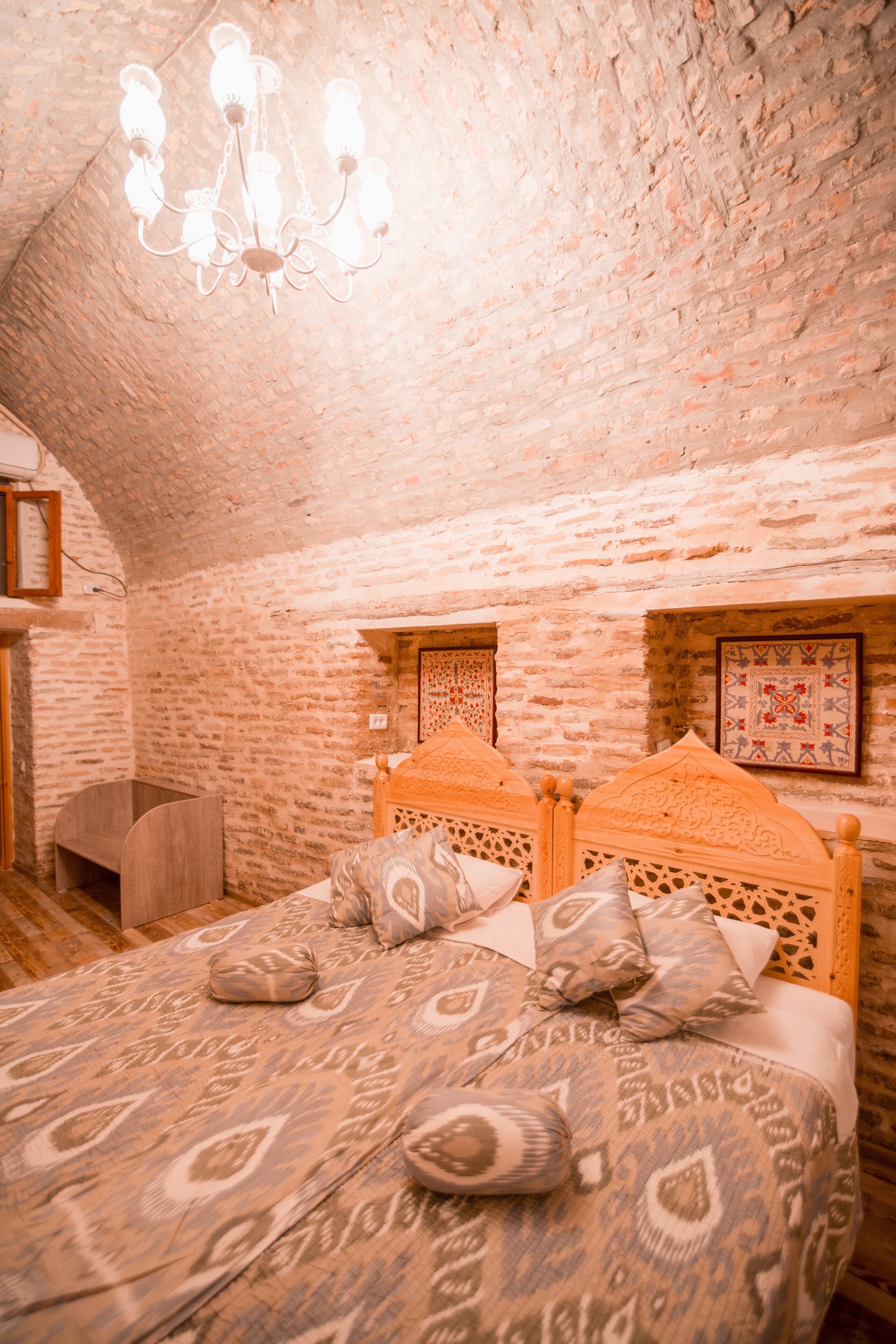 Une chambre avec un mur en briques, un lit en bois avec une literie à motifs, un lustre et un petit banc en bois.