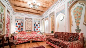 Une pièce avec un plafond en bois, des décorations murales élaborées, un lit avec une literie rouge, un canapé recouvert d’un tissu à motifs et un plancher en bois.