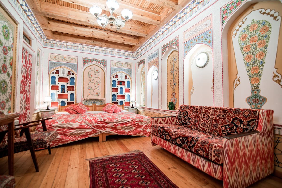 Ein Zimmer mit Holzboden, einer Holzbalkendecke, aufwändigen Wandverzierungen, einem Bett mit roter Bettwäsche und einem Sofa mit gemustertem Stoff.