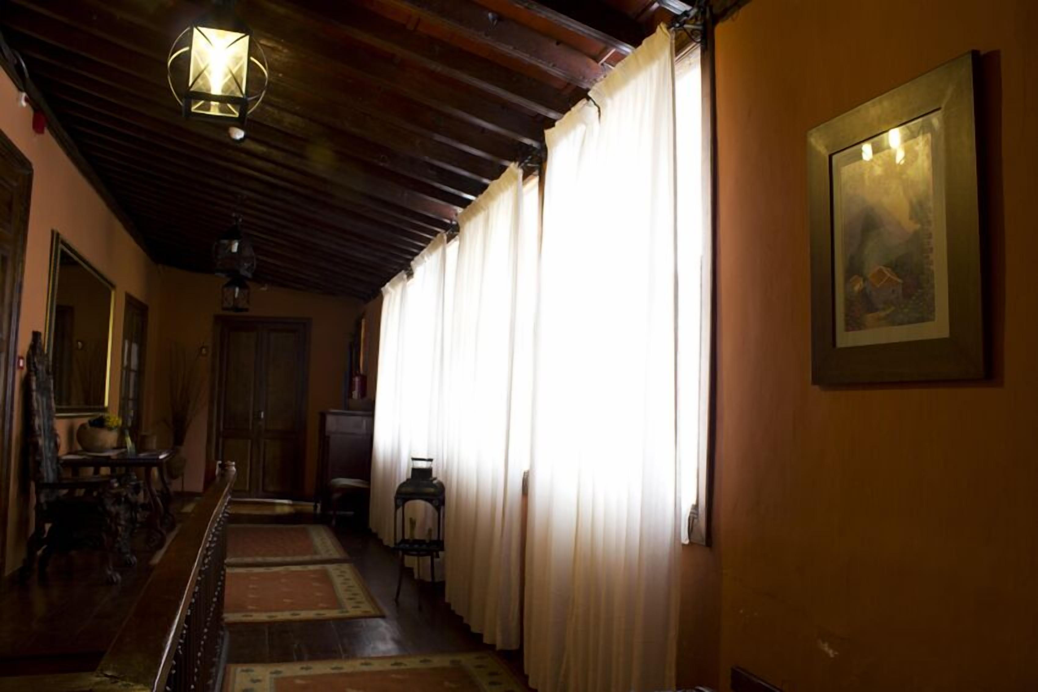 A hallway with a wooden ceiling, a chandelier, a large window with curtains, a framed picture on the wall, and a small table with a lamp.