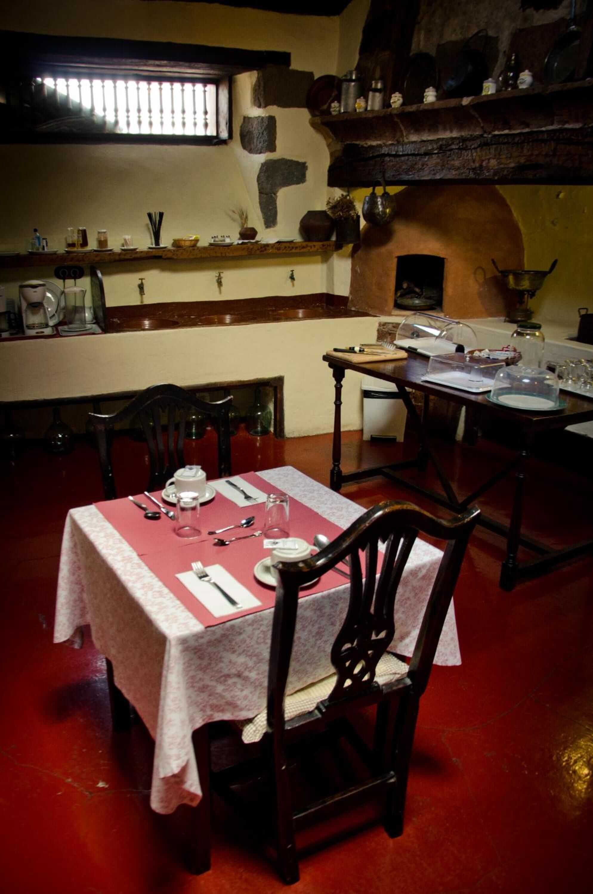 Comedor con mesa dispuesta para cuatro, una silla de madera y zona de cocina con estufa y estanterías arriba.
