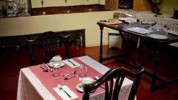 Comedor con mesa dispuesta para cuatro, una silla de madera y zona de cocina con estufa y estanterías arriba.