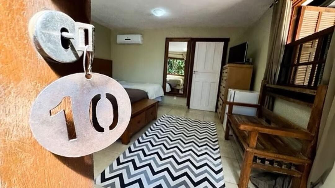 A room with a bed, a desk, a chair, a door with a number plate, and a patterned rug.
