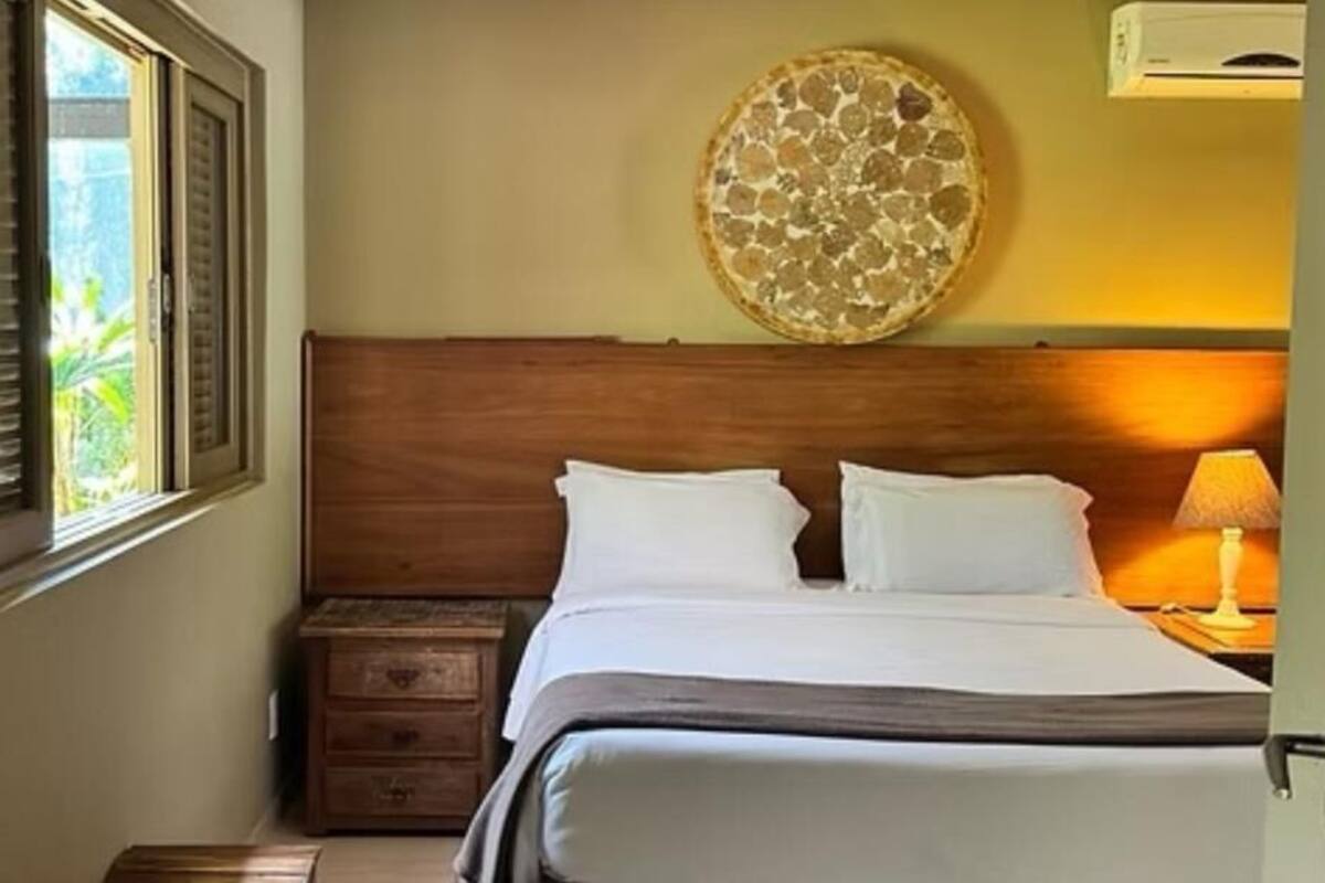 Un dormitorio con cabecera de madera, cama con ropa blanca, una obra de arte en la pared, una mesita de noche y una ventana con persianas.
