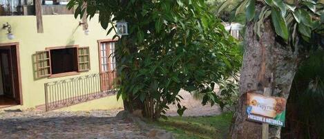 Un sendero adoquinado conduce a un edificio amarillo con una ventana y una puerta, rodeado de abundante vegetación y un letrero que dice