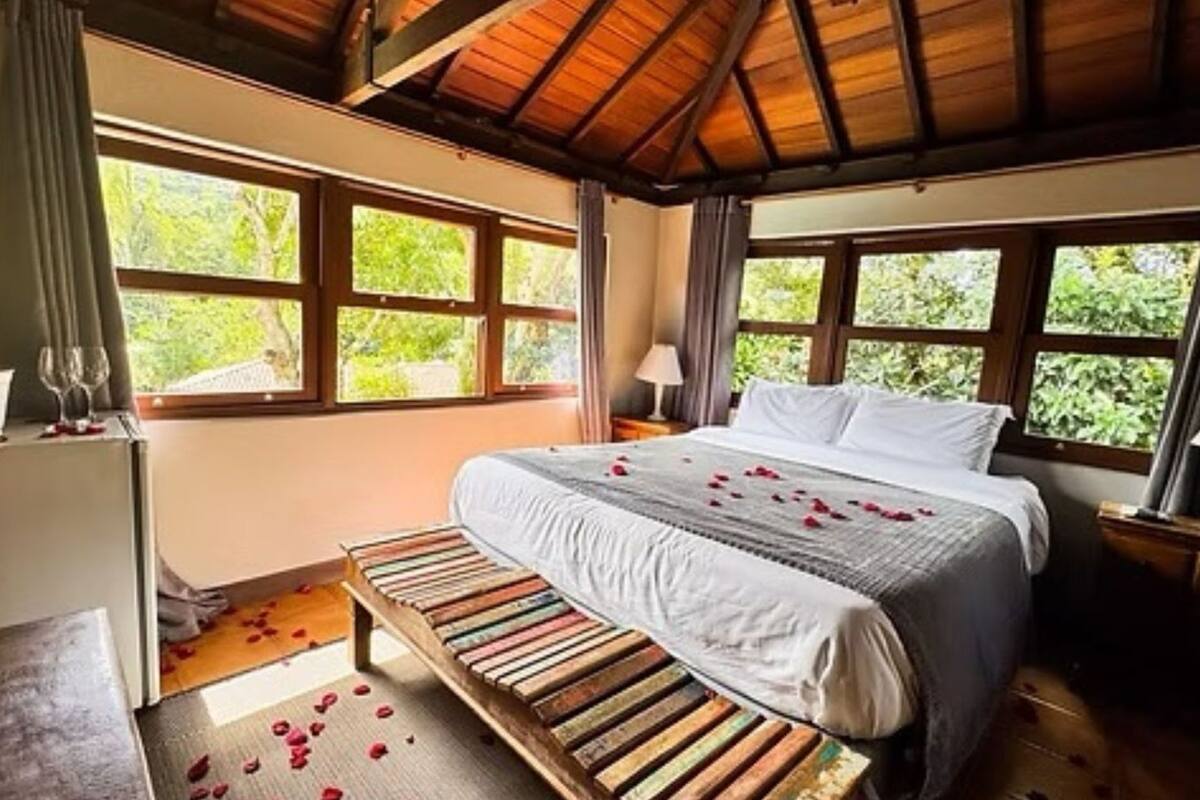 Un dormitorio con un banco de madera, una cama con ropa de cama blanca y ventanas grandes con cortinas.