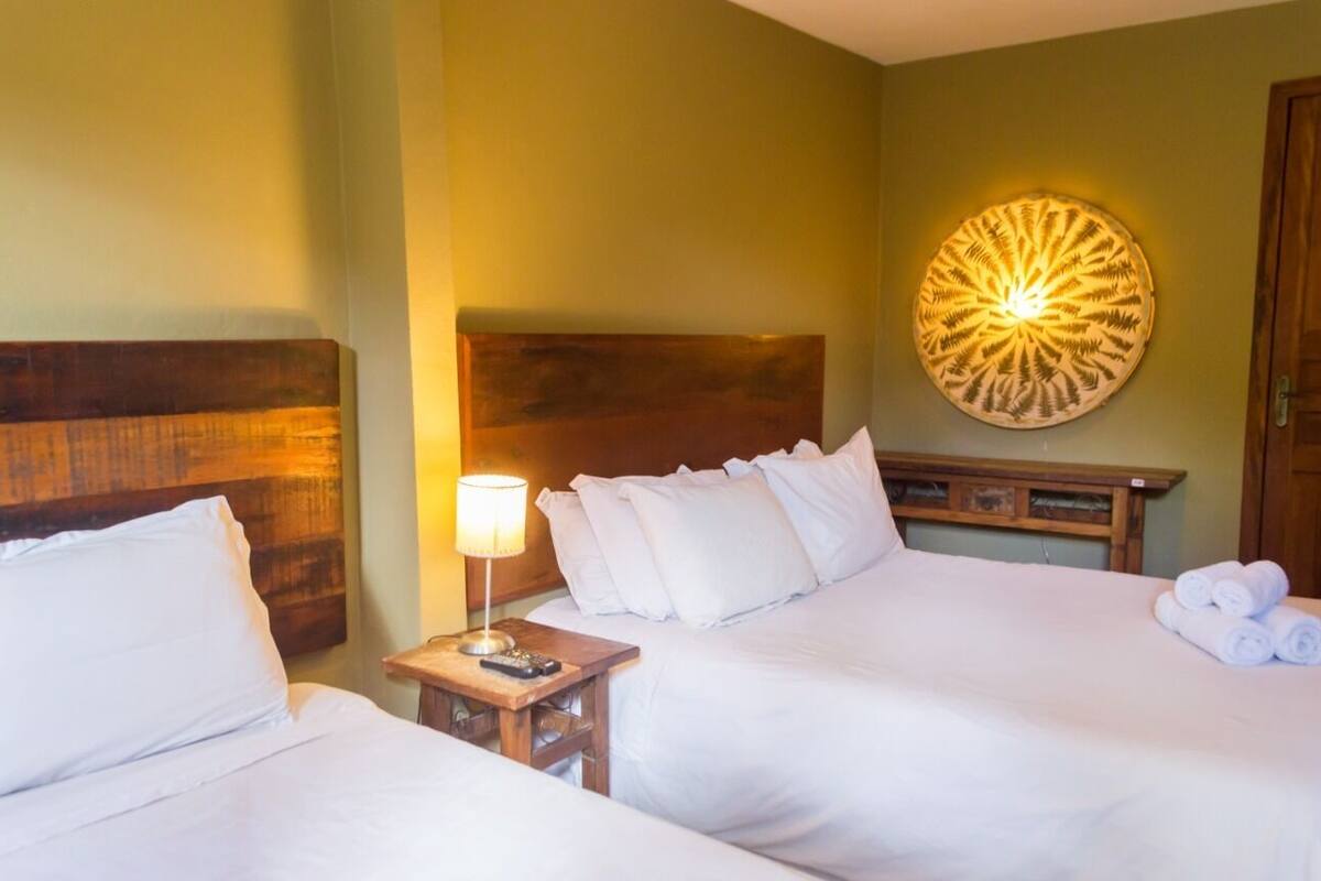 Habitación de hotel con dos camas, cabecero de madera, lámpara de pared y mesita de noche con lámpara y control remoto.