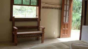 Una habitación con una cama, un banco de madera, una ventana y una puerta abierta que da al exterior.