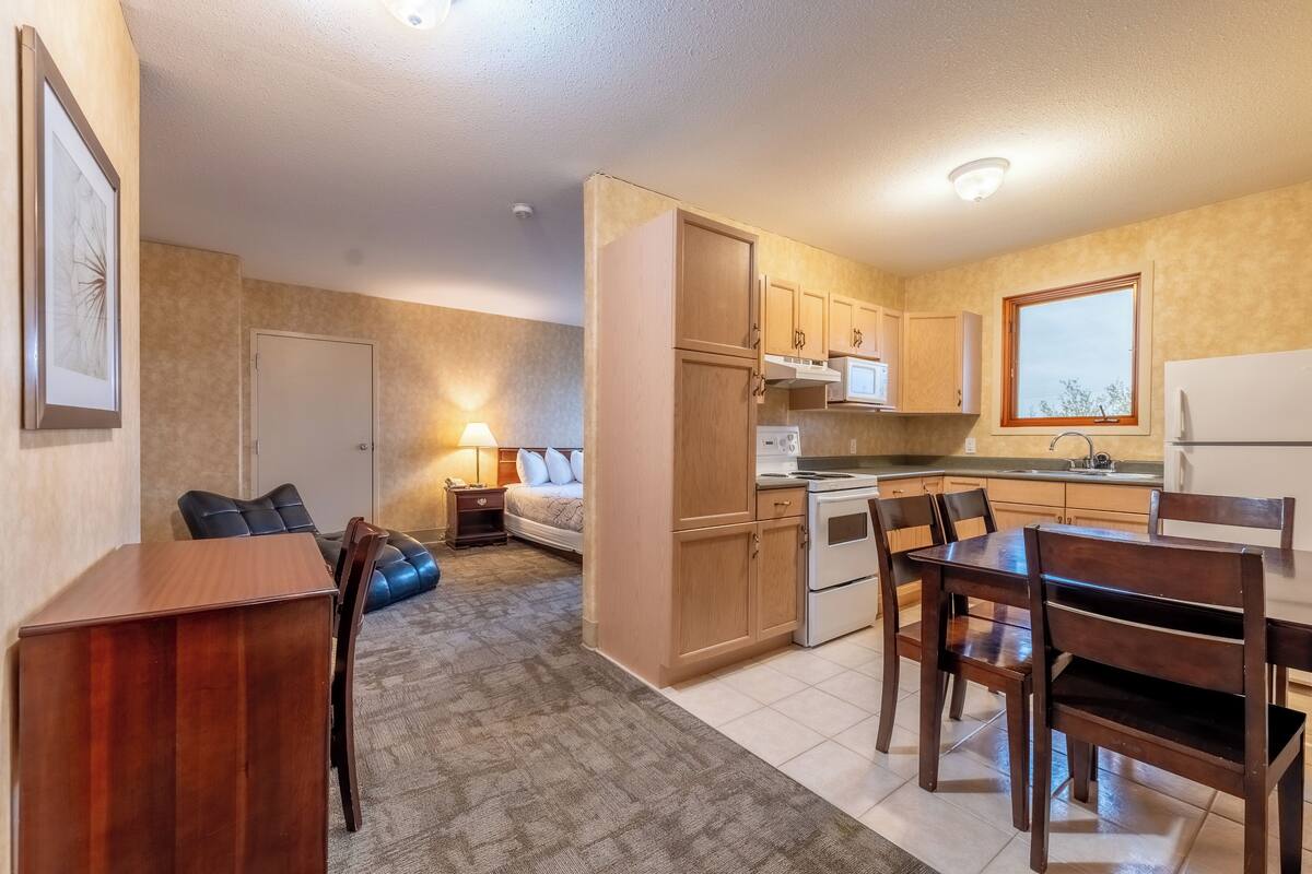 A compact kitchen with wooden cabinets, a microwave, and a refrigerator. There is a dining area with a table and chairs, and a bedroom with a bed and a nightstand.