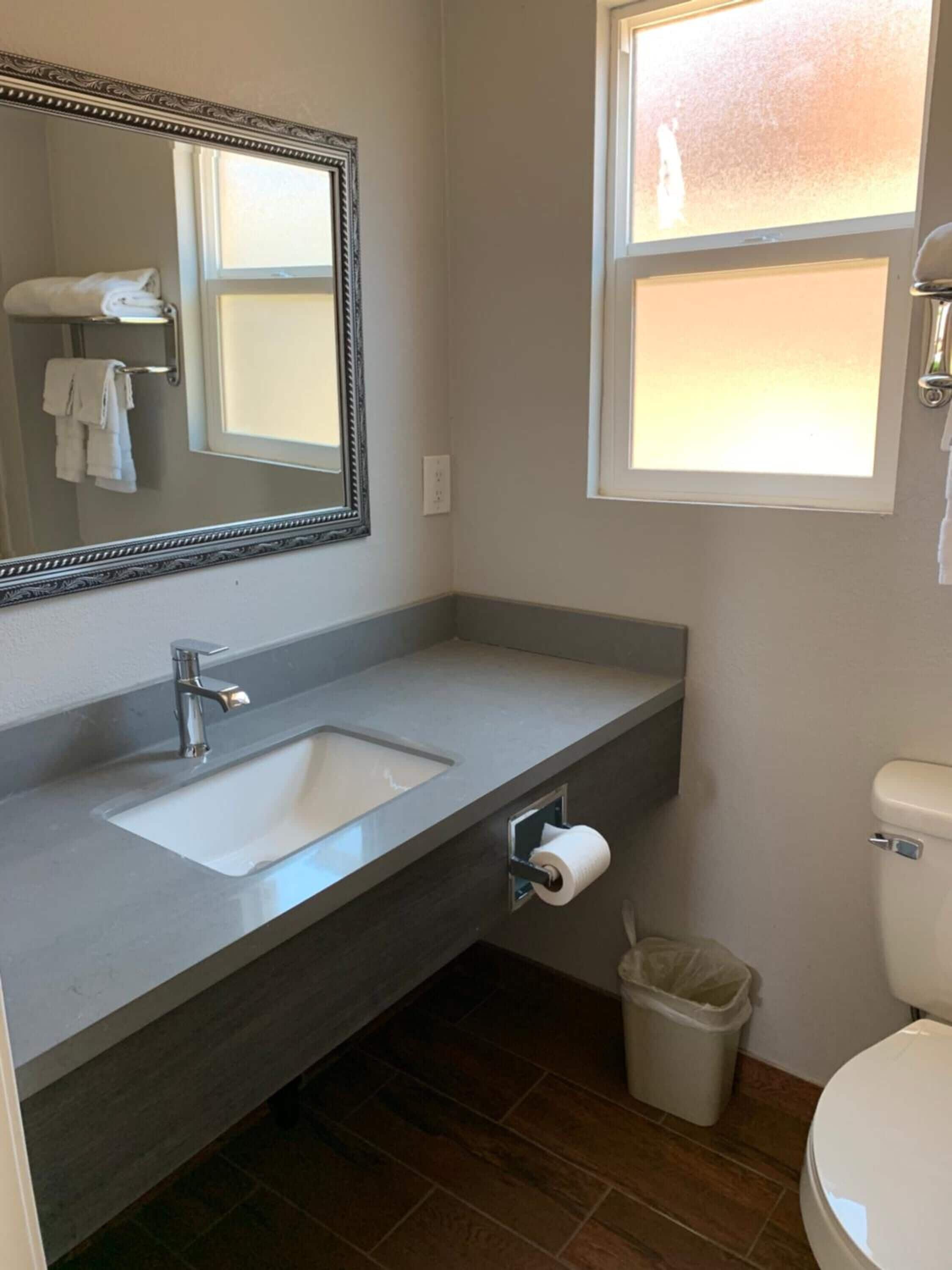 A bathroom with a double sink, a toilet, a trash can, and a window.