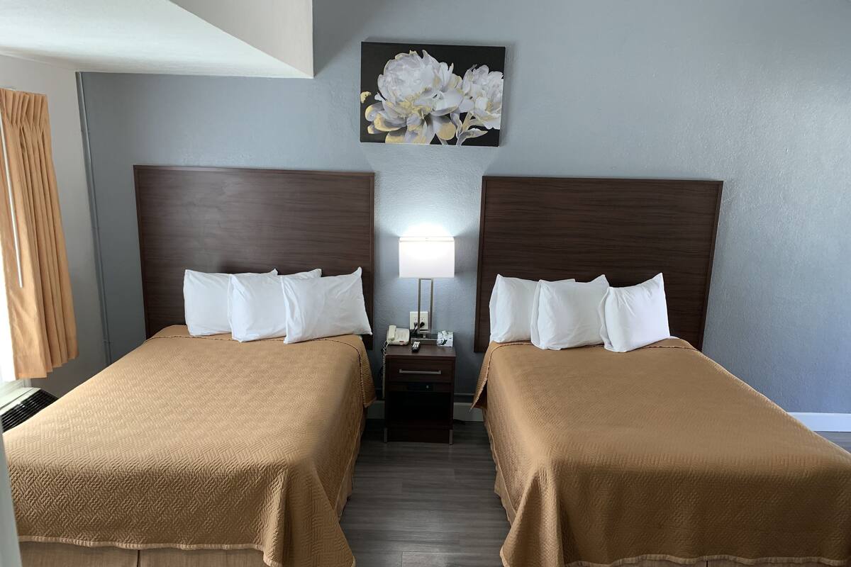 A hotel room with two beds, beige bedding, a nightstand with a lamp, and a floral painting on the wall.