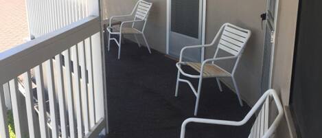A balcony with white metal chairs and a small table, a door with a screen, and a view of the outside.