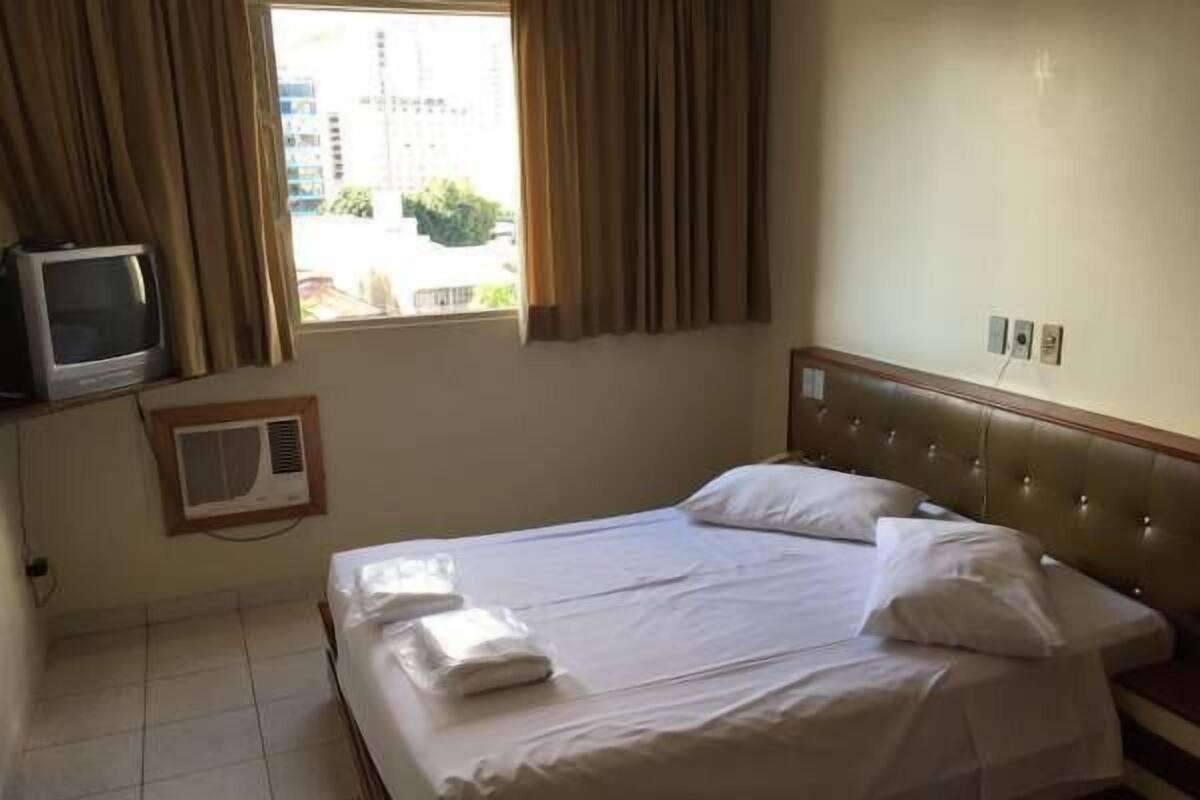 A hotel room with a bed, a television, a window with curtains, and a thermostat on the wall.