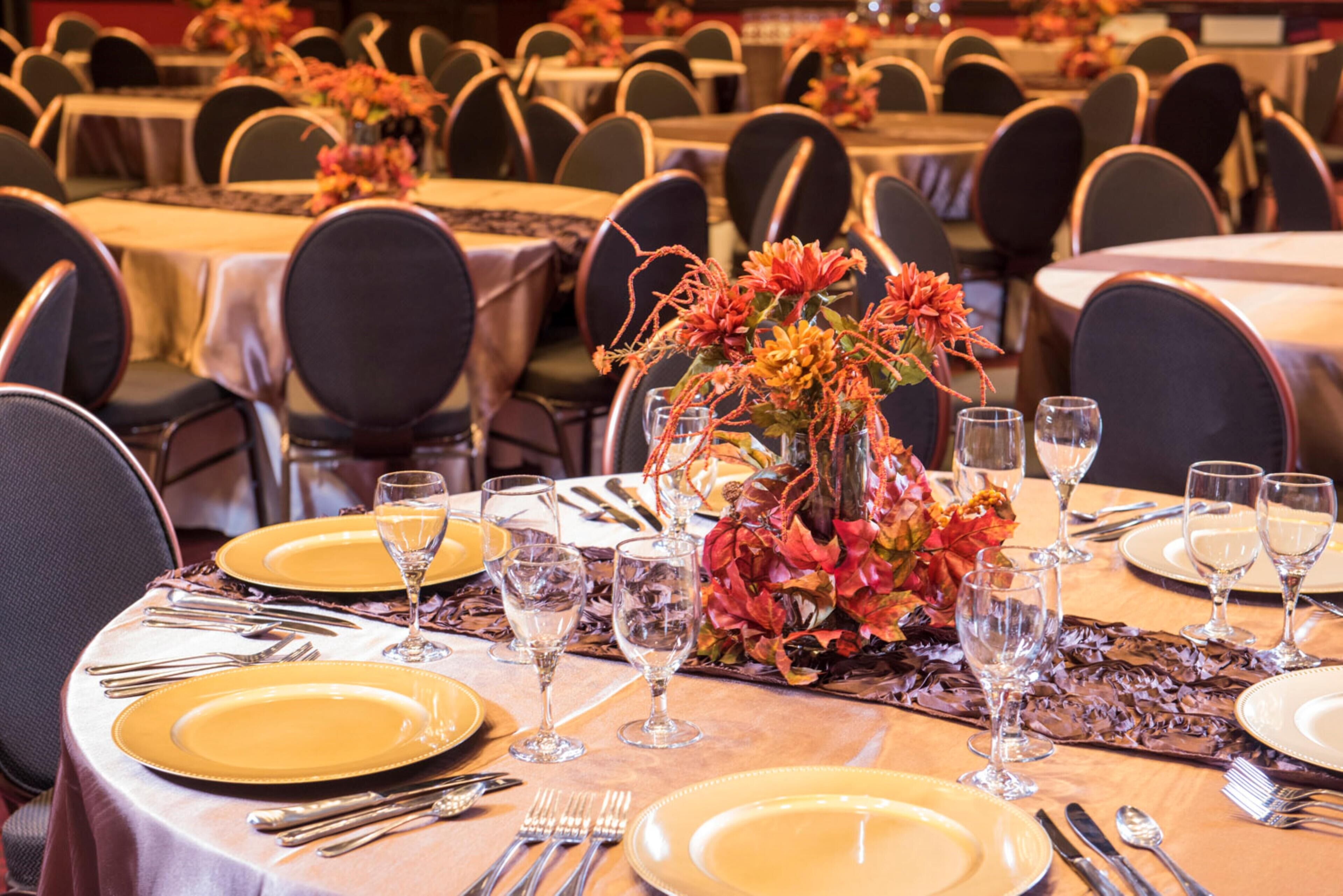 Uma sala de banquetes com mesas redondas preparadas para refeições, cada uma com pratos amarelos, talheres e copos de vinho.