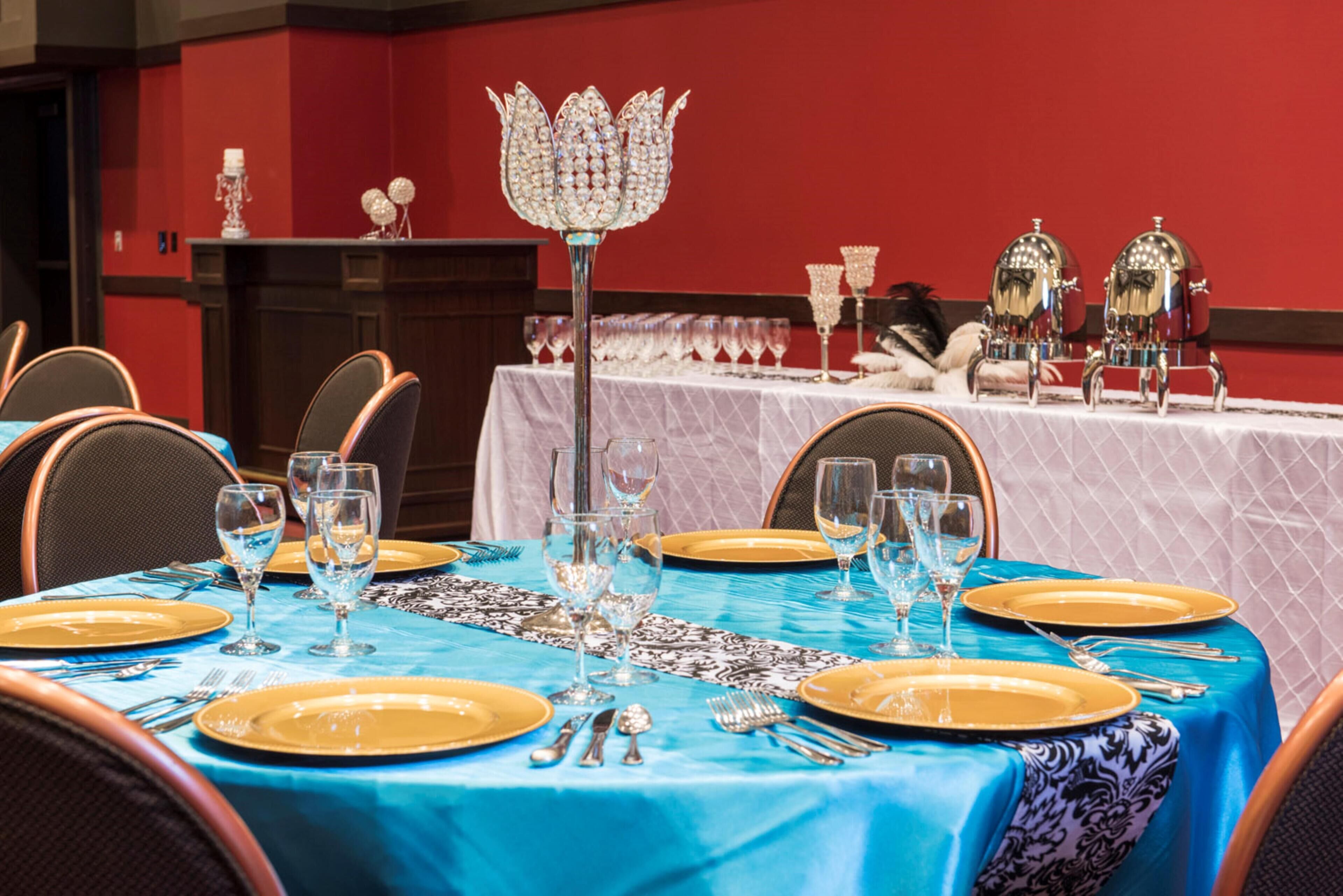 Una mesa de comedor dispuesta con platos dorados, cubiertos de plata y cristalería, con mantel azul y una pieza decorativa en el centro.