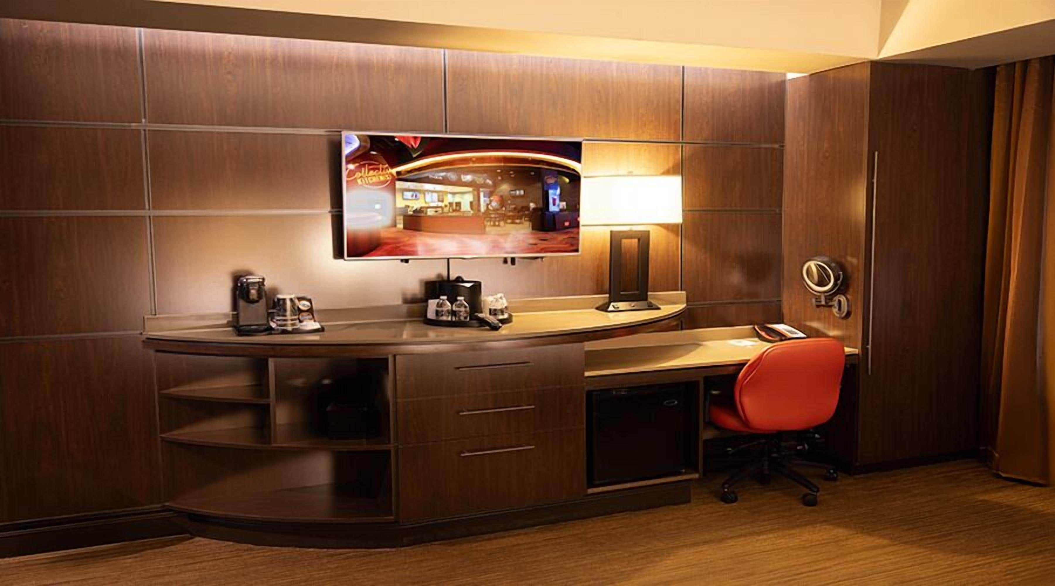 Ett hotellrum med ett böjt skrivbord, en platt-TV som är monterad på väggen, en skrivbordslampa, en röd kontorsstol och en minibar.