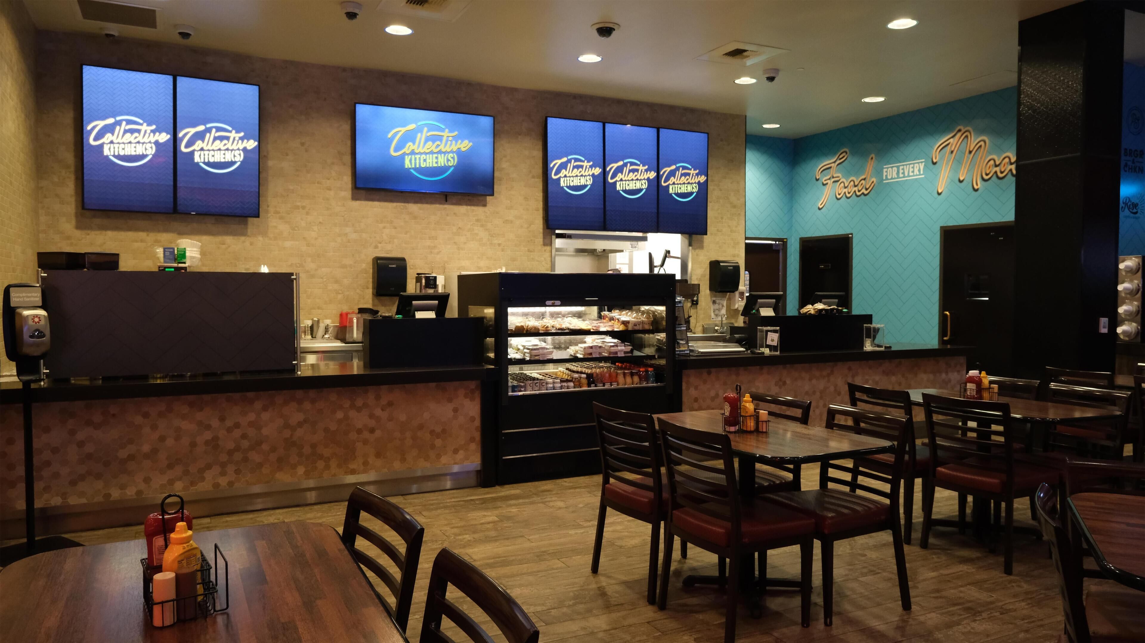 A restaurant interior with multiple tables and chairs, a counter with a refrigerated display case, and wall-mounted screens displaying the restaurant's name.