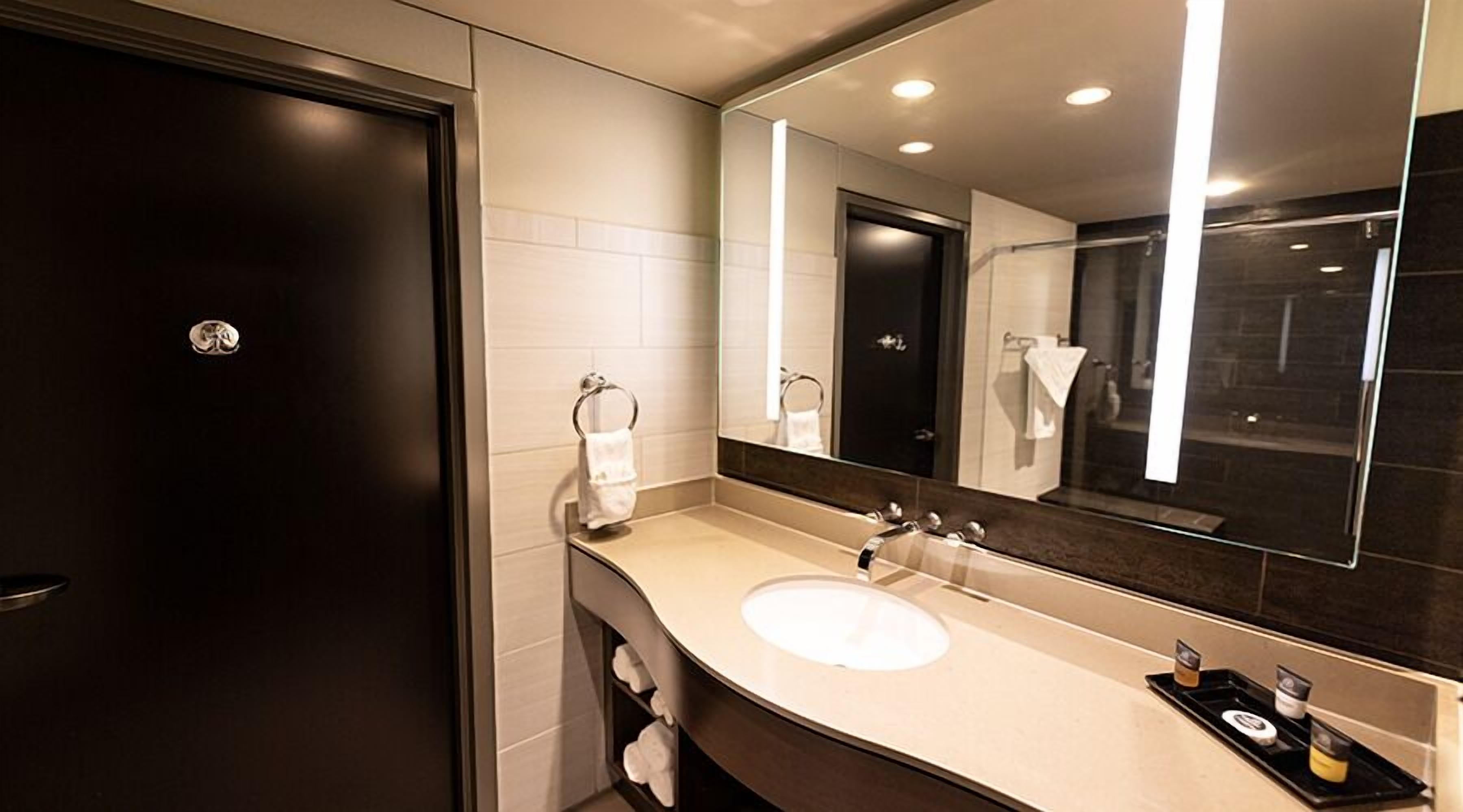 Un baño moderno con una puerta oscura, un lavabo blanco, un espejo grande y una bandeja con artículos de aseo.