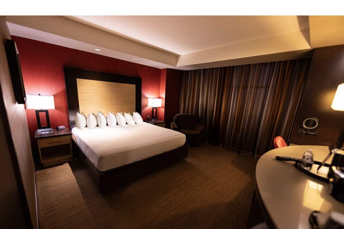 A hotel room with a large bed, two bedside tables with lamps, a chair, a desk with a chair, and a curtain.