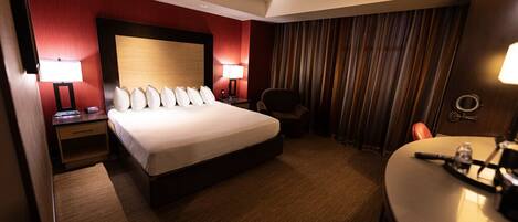 Ein Hotelzimmer mit einem großen Bett, zwei Nachttischen mit Lampen, einem Sessel, einem Schreibtisch mit Stuhl und einem Vorhang.