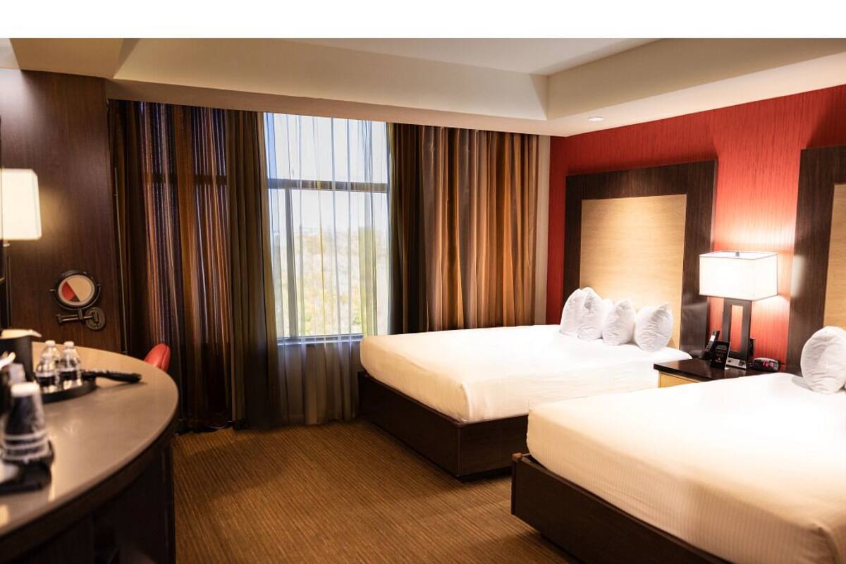 A hotel room with two beds, a desk, a chair, a lamp, and a window with curtains.