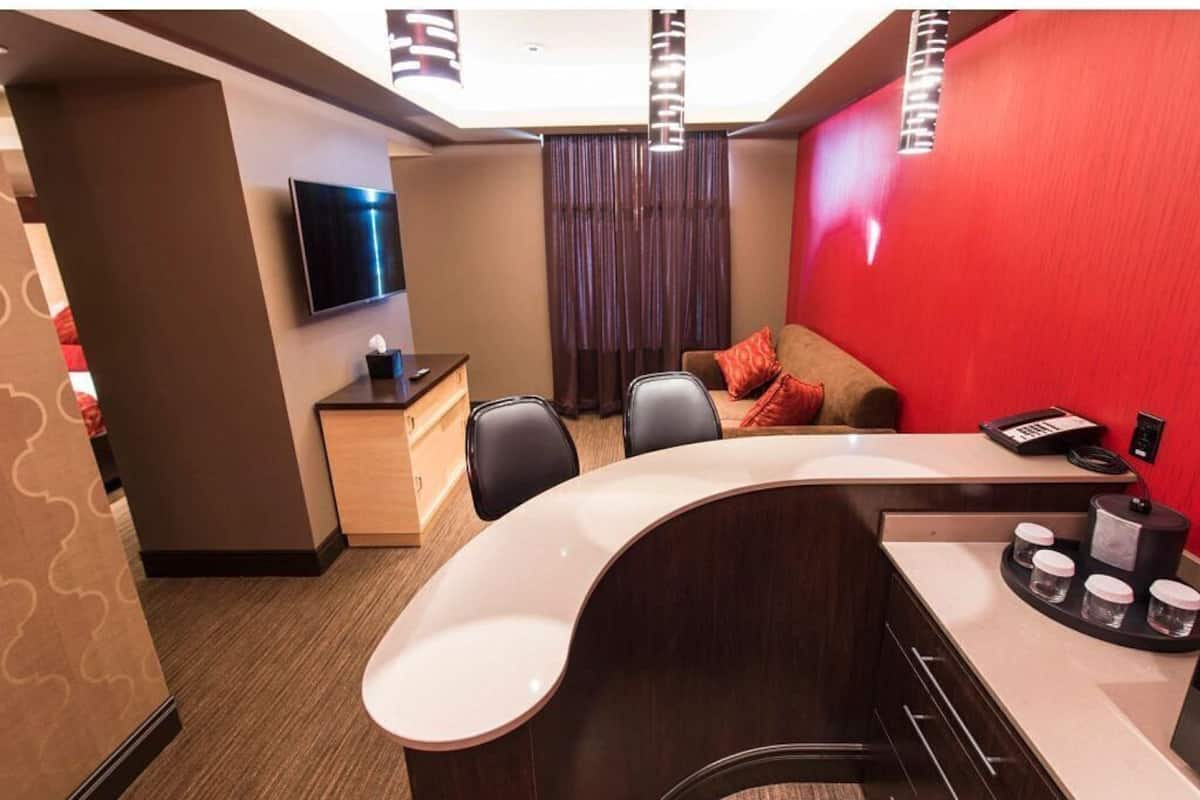 A modern hotel room with a curved desk, a flat-screen TV, a small wooden side table, and a red accent wall.