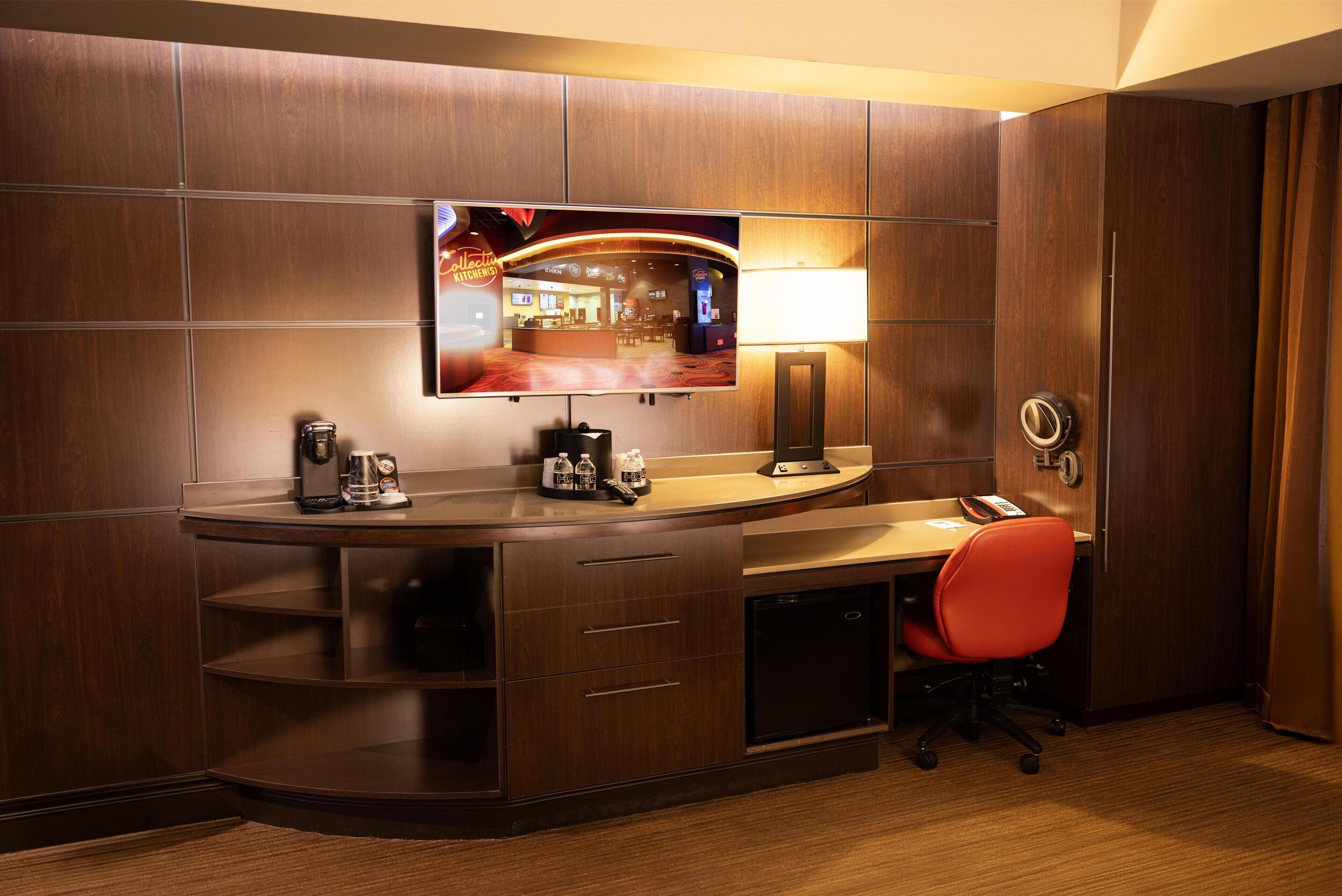A hotel room with a curved desk, a flat-screen TV mounted on the wall, a desk lamp, a red office chair, and a minibar.