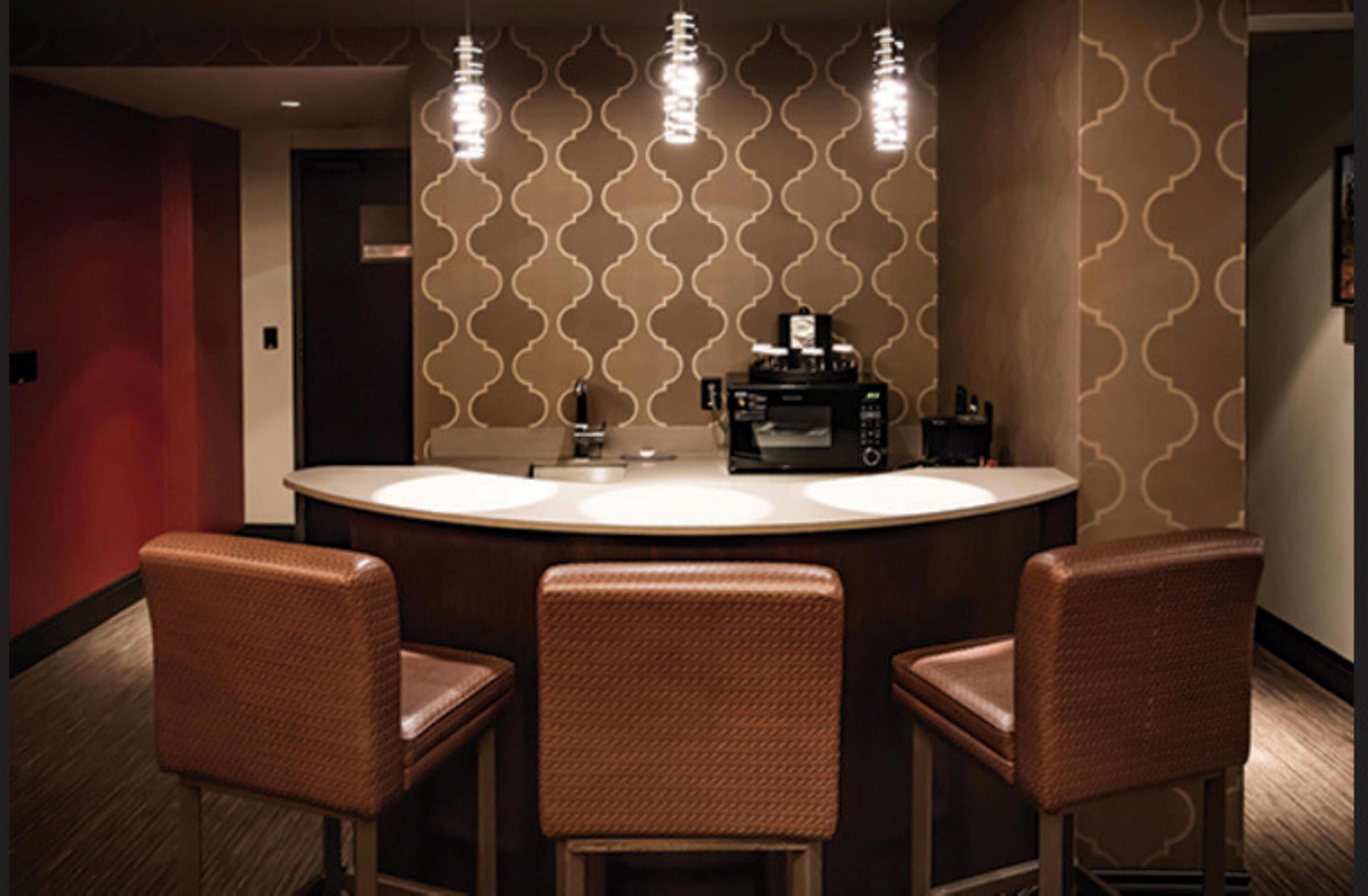 Une salle de bain d’hôtel avec un lavabo rond, trois chaises marron et un four à micro-ondes posé sur le comptoir.