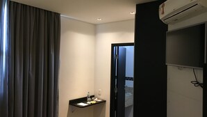 Una habitación de hotel con una cortina oscura, un estante pequeño con artículos de aseo, un equipo de aire acondicionado y una puerta que da a otra habitación.