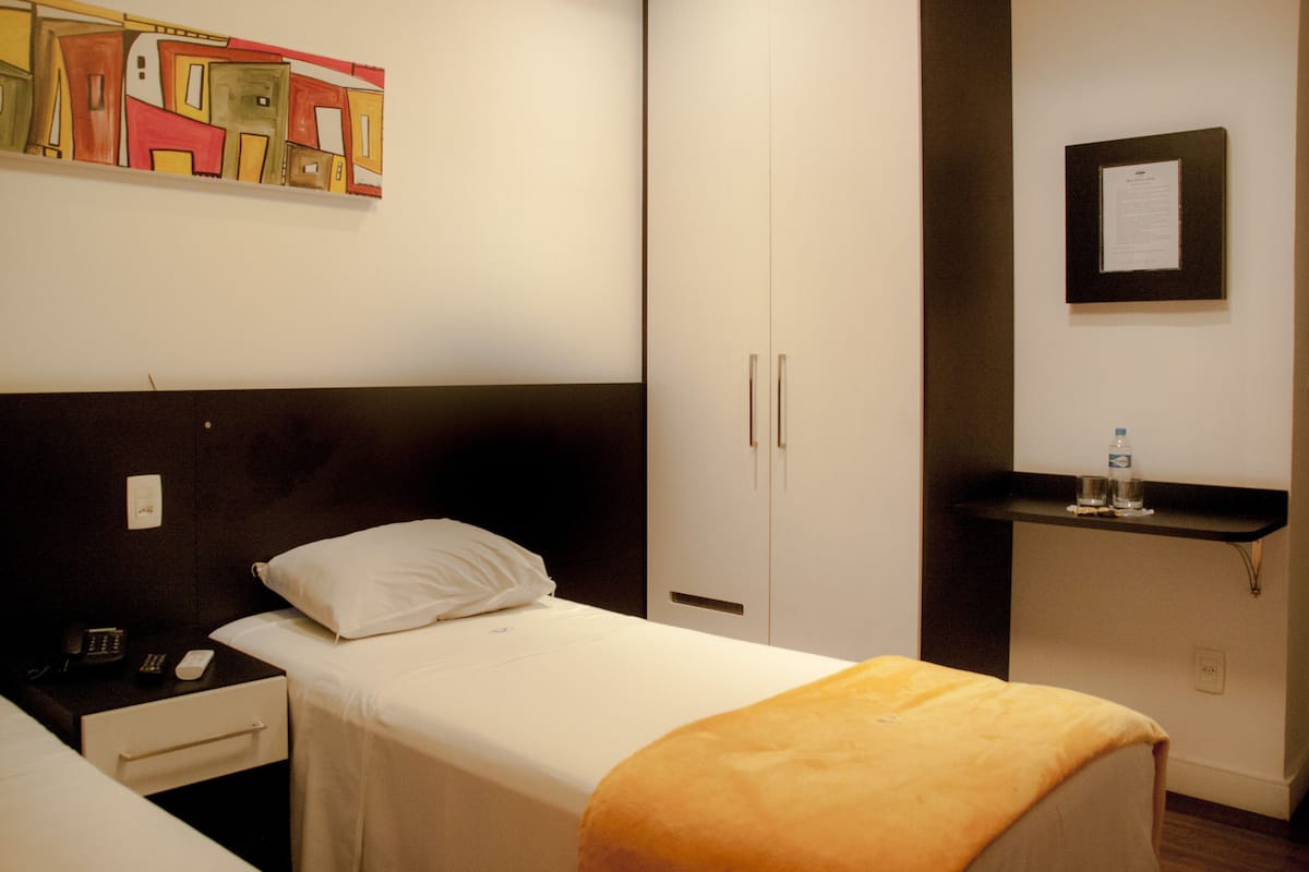 Habitación de hotel con una cama individual, una mesita de noche con un teléfono, una pequeña mesa con una botella de agua y un certificado enmarcado en la pared.