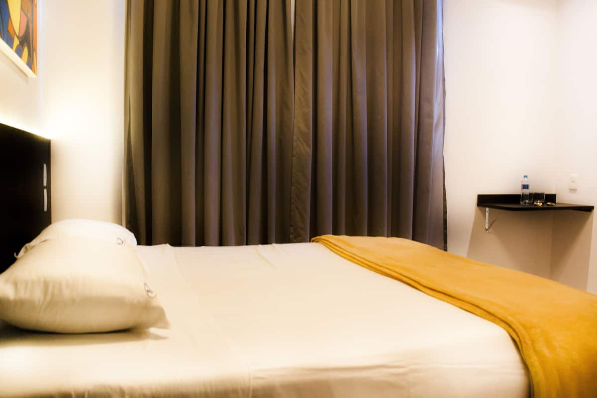 Una habitación de hotel con una cama, cabecero, mesita de noche y cortina.