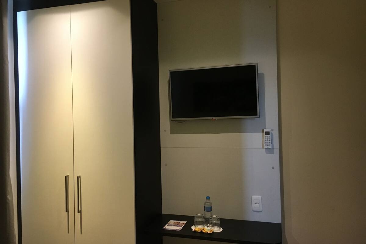 Habitación de hotel con un armario cerrado, un televisor montado, una mesita con una botella de agua, vasos y una revista.