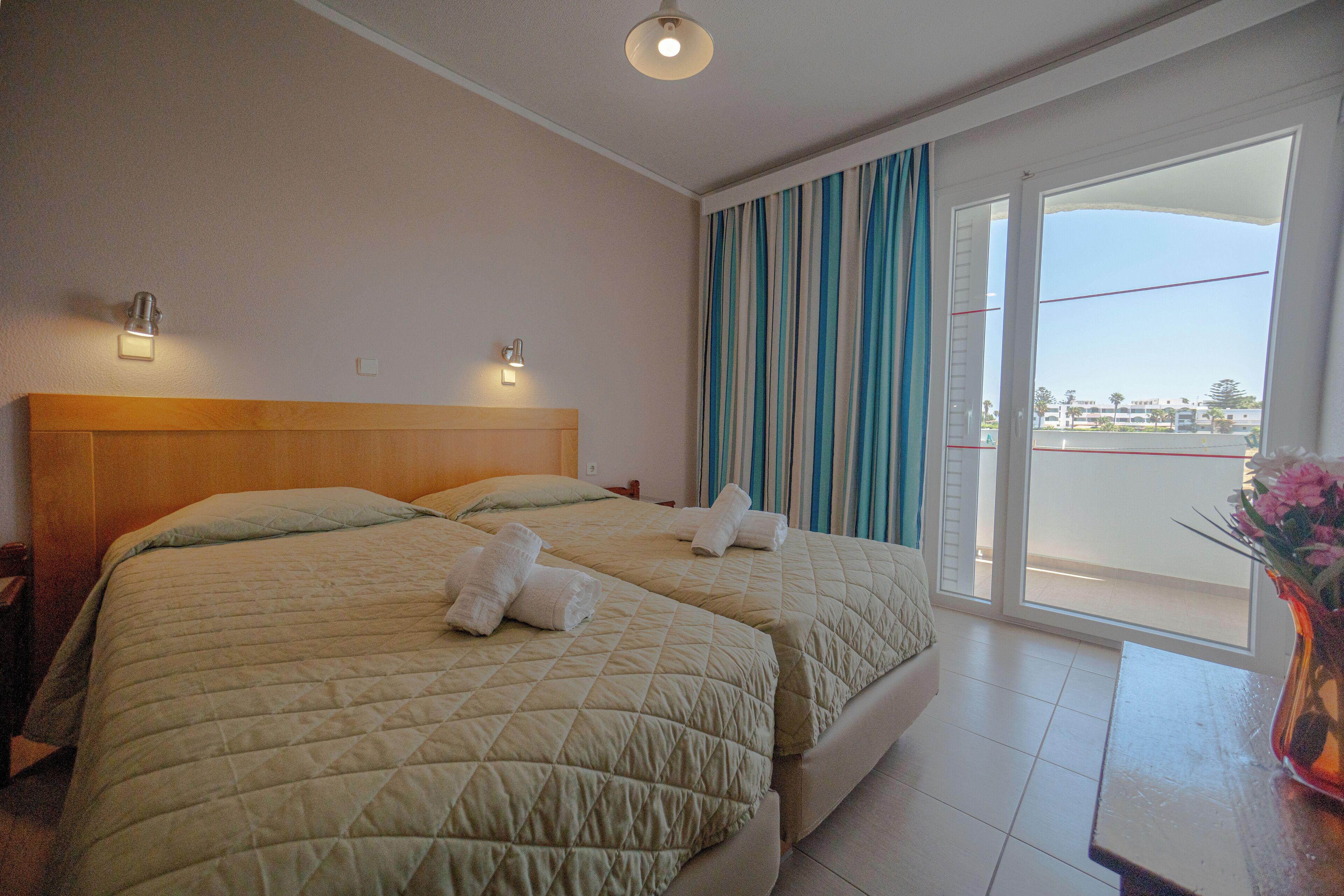 Een hotelkamer met twee bedden, een houten hoofdbord, een schuifdeur naar het balkon en een bloemenvaas op het nachtkastje.