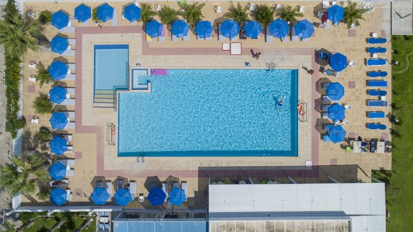 An aerial view of a swimming pool area with multiple sun loungers and umbrellas, surrounded by palm trees and a tiled walkway.