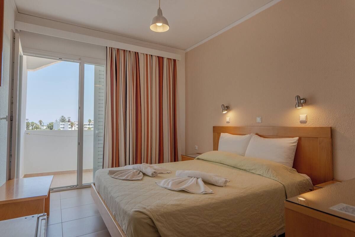 A hotel room with a bed, a nightstand, a desk, and a view through a sliding glass door.