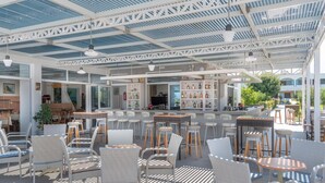 An outdoor seating area with wicker chairs and tables, a bar with shelves of bottles, and a covered ceiling with a grid pattern. - Tropical Sol (Kos)