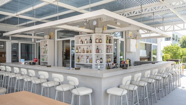 A modern bar area with a long counter, white bar stools, and shelves stocked with bottles. - Tropical Sol (Kos)