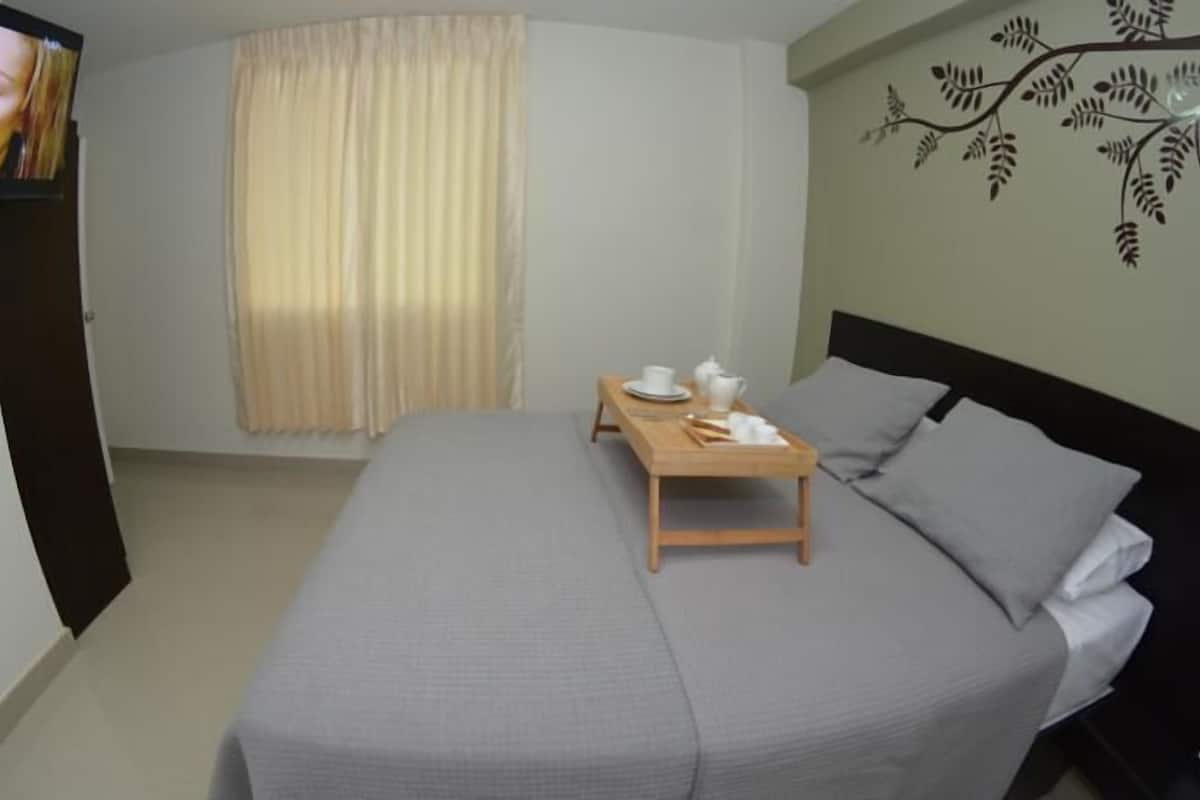 Habitación de hotel con cama, una mesita con juego de té, televisor en la pared y ventana con cortinas.