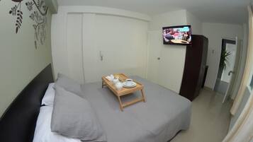 Una habitación de hotel con una cama, una pequeña mesa de madera con una taza y un platillo, un televisor en la pared y una puerta con una planta a su lado.