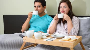 Um casal sentado na cama com uma mesinha entre eles, segurando xícaras de café.