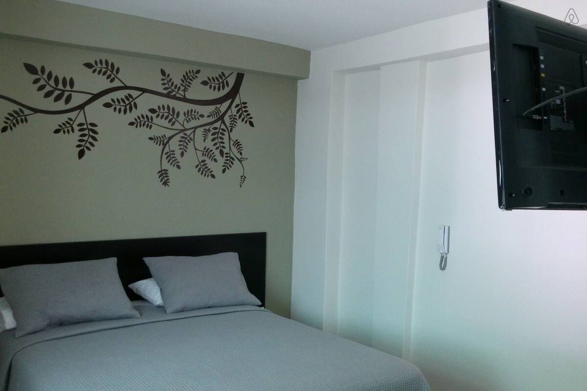 Un dormitorio con cama, televisión instalada en la pared y un diseño decorativo con hojas en la pared.