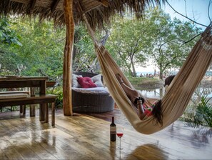 A hammock chair with a cushion, a wooden table, and a bottle of wine on a wooden deck with a thatched roof.