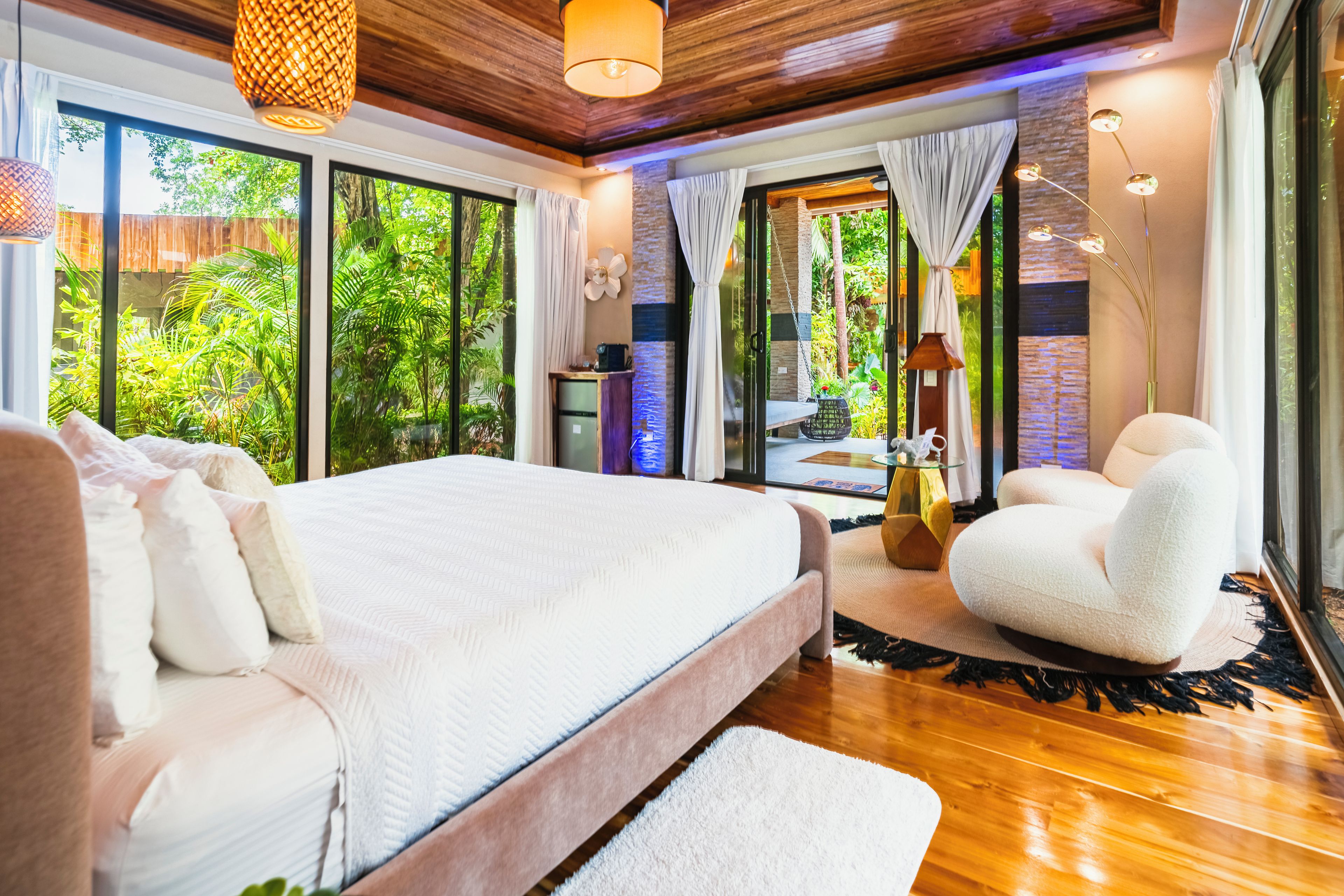 A bedroom with a large bed, a sitting area with a chair, and a view of greenery through the windows.